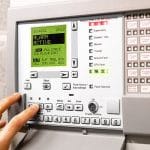 Person pressing a button on a business alarm system control panel. AirDial provides an all-in-one POTS line replacement for old emergency systems during the copper transition.